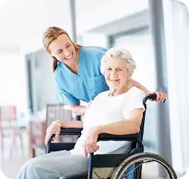 Smiling nurse in teal scrubs standing behind an older woman in a wheelchair