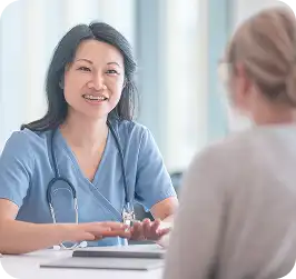 Nurse in scrubs with a stethoscope smiling and talking with a patient across a desk